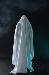 Photo of Creepy ghost. Man covered with white sheet in smoke on black background