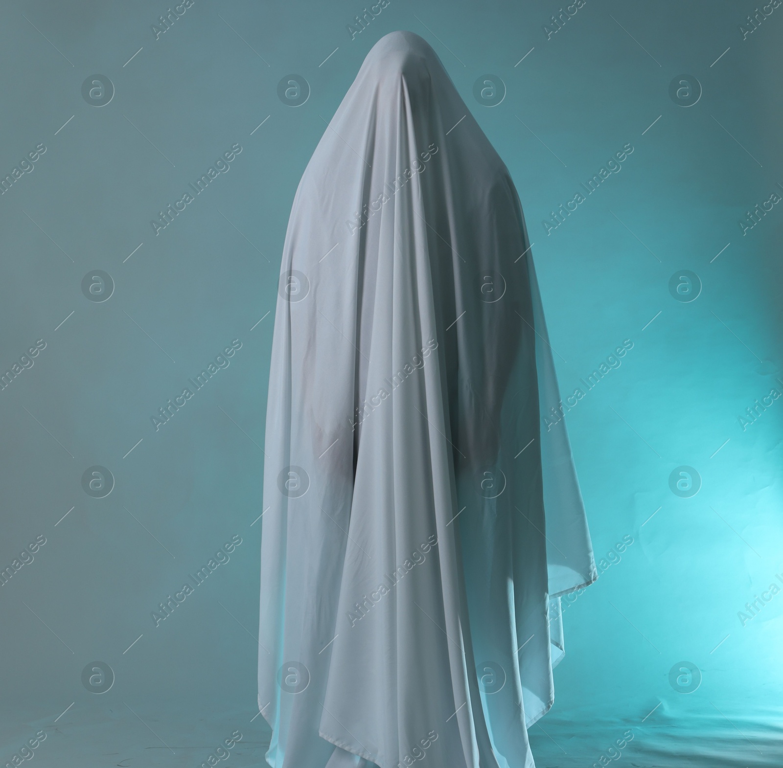 Creepy ghost. Man covered with white sheet in smoke on light blue background Photo of Creepy ghost. Man covered with white sheet in smoke on light blue background