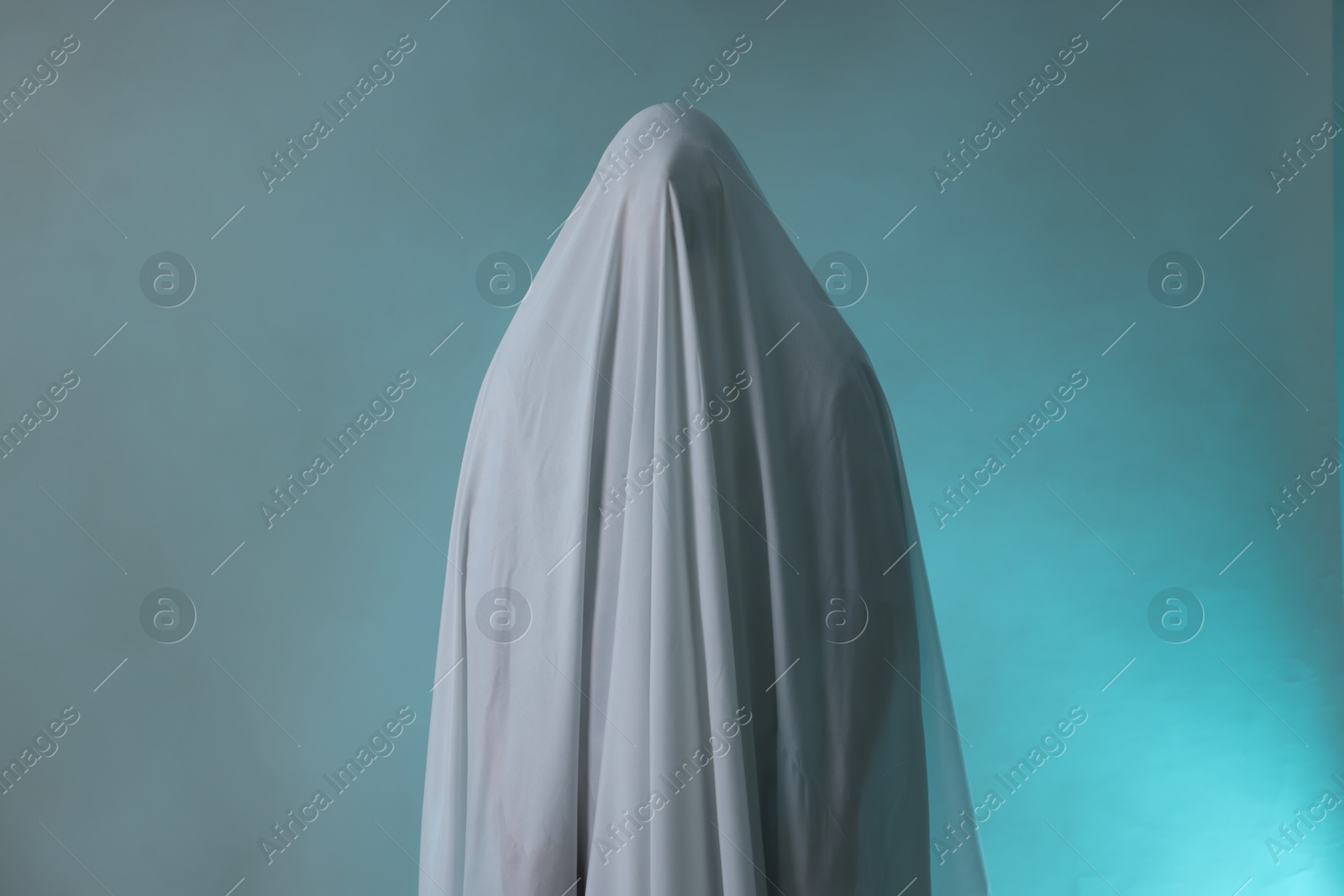 Photo of Creepy ghost. Man covered with white sheet in smoke on light blue background