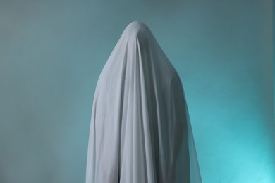 Photo of Creepy ghost. Man covered with white sheet in smoke on light blue background