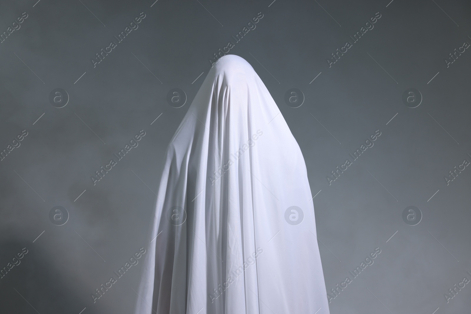 Photo of Creepy ghost. Man covered with white sheet in smoke on light grey background