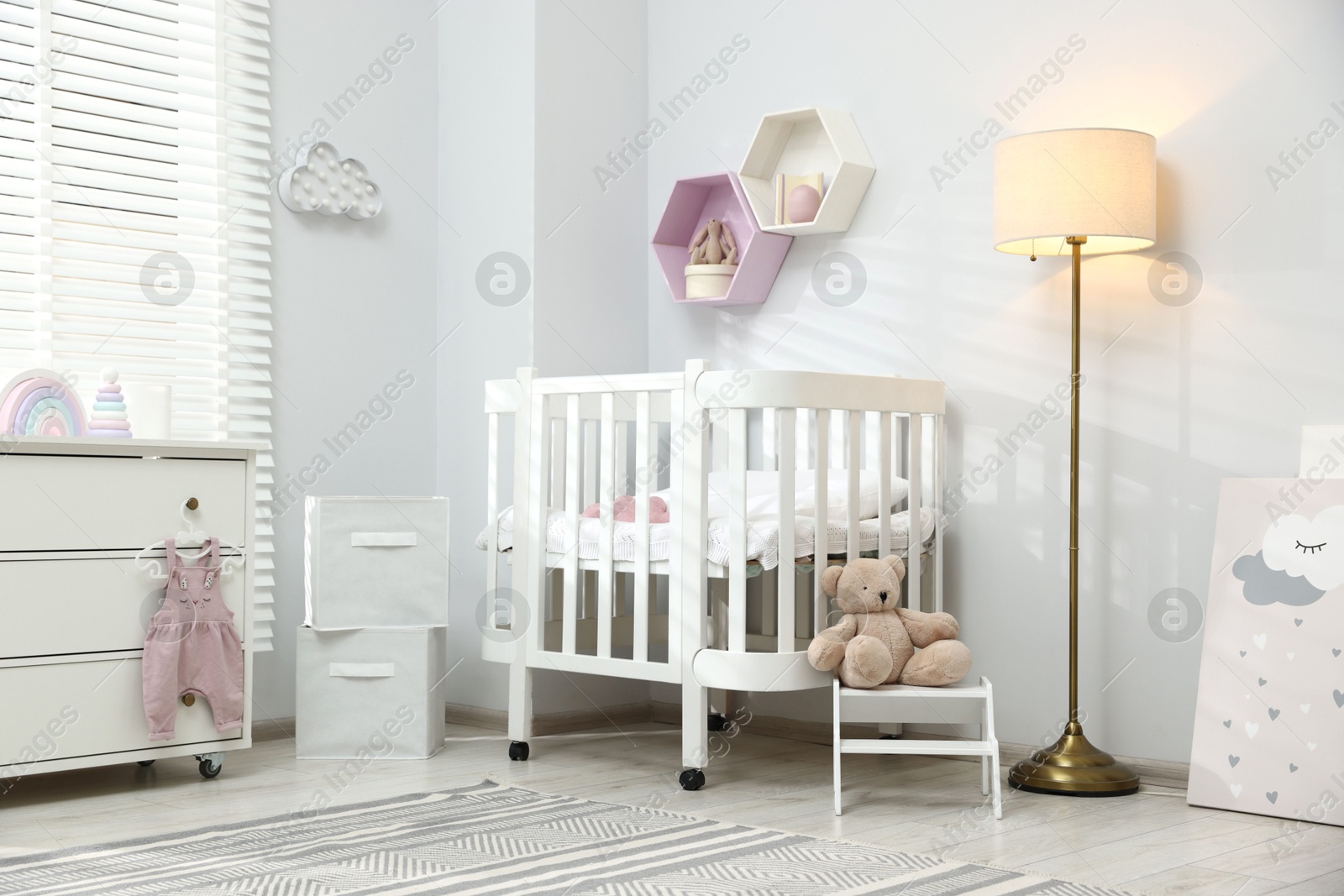 Stylish white crib, chest of drawers, lamp, toys and decor elements in baby's room. Interior design Photo of Stylish white crib, chest of drawers, lamp, toys and decor elements in baby's room. Interior design