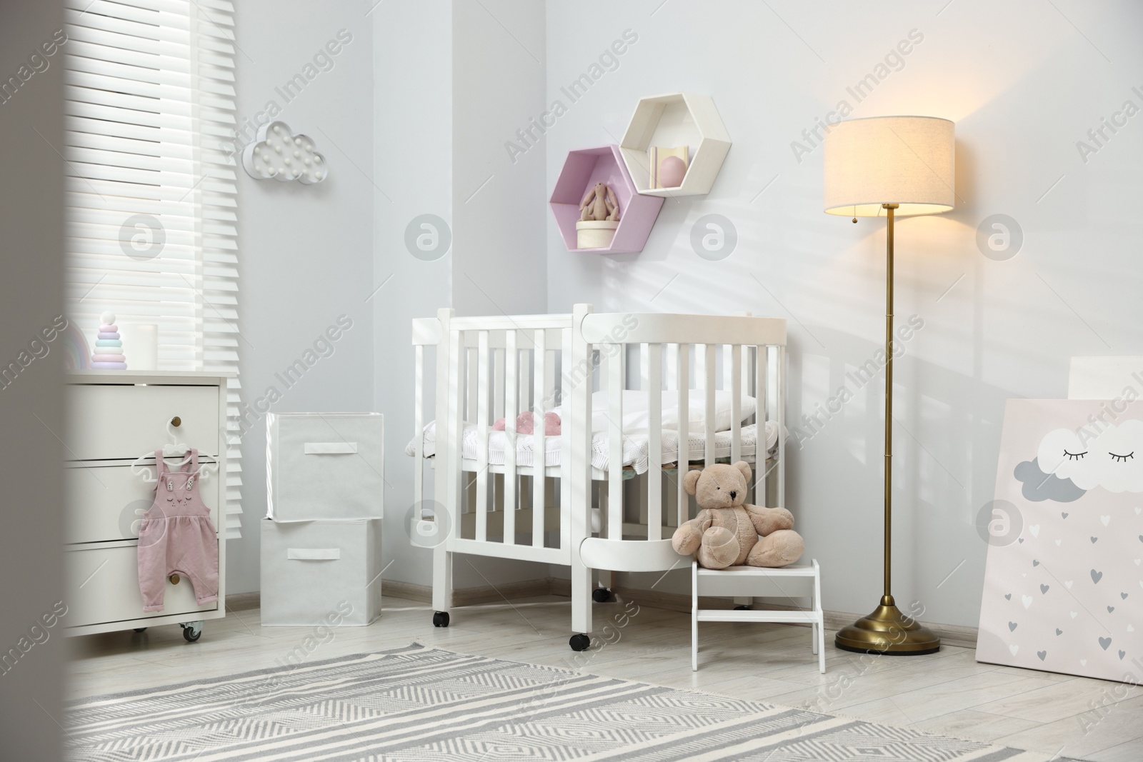 Stylish white crib, chest of drawers, lamp, toys and decor elements in baby's room. Interior design Photo of Stylish white crib, chest of drawers, lamp, toys and decor elements in baby's room. Interior design