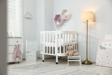 Photo of Stylish white crib, chest of drawers, lamp, toys and decor elements in baby's room. Interior design