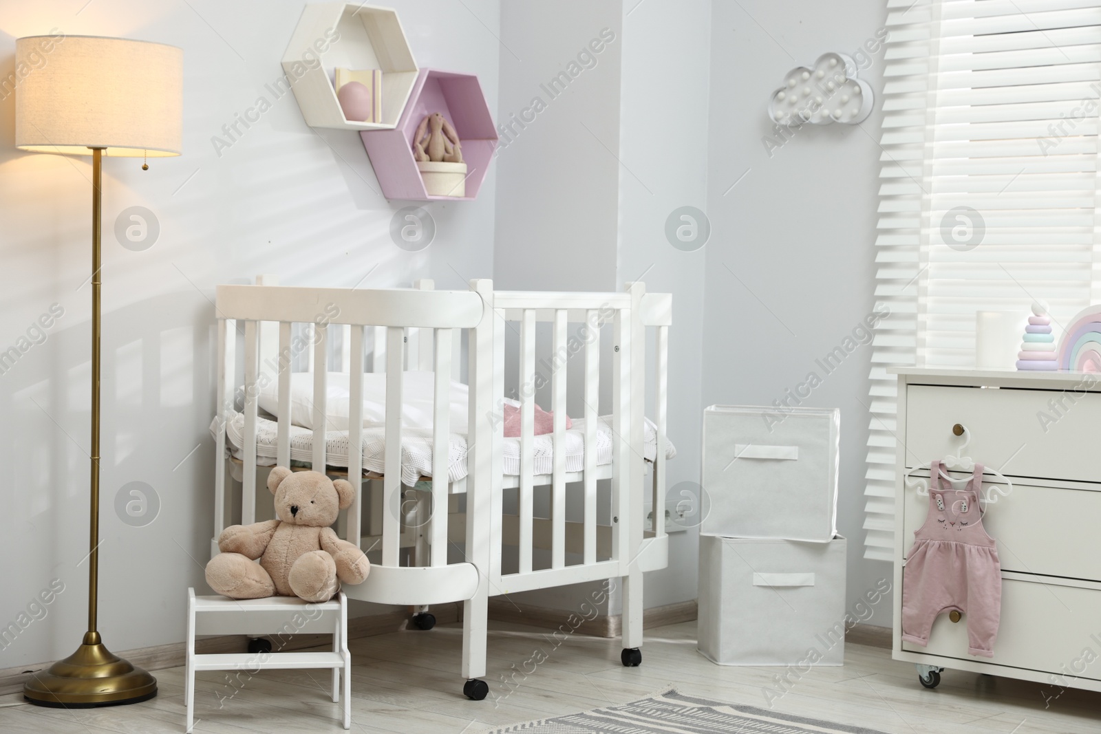Stylish white crib, chest of drawers, lamp, toys and decor elements in baby's room. Interior design Photo of Stylish white crib, chest of drawers, lamp, toys and decor elements in baby's room. Interior design