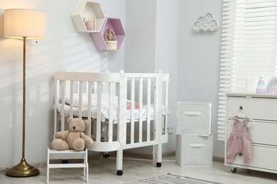 Photo of Stylish white crib, chest of drawers, lamp, toys and decor elements in baby's room. Interior design