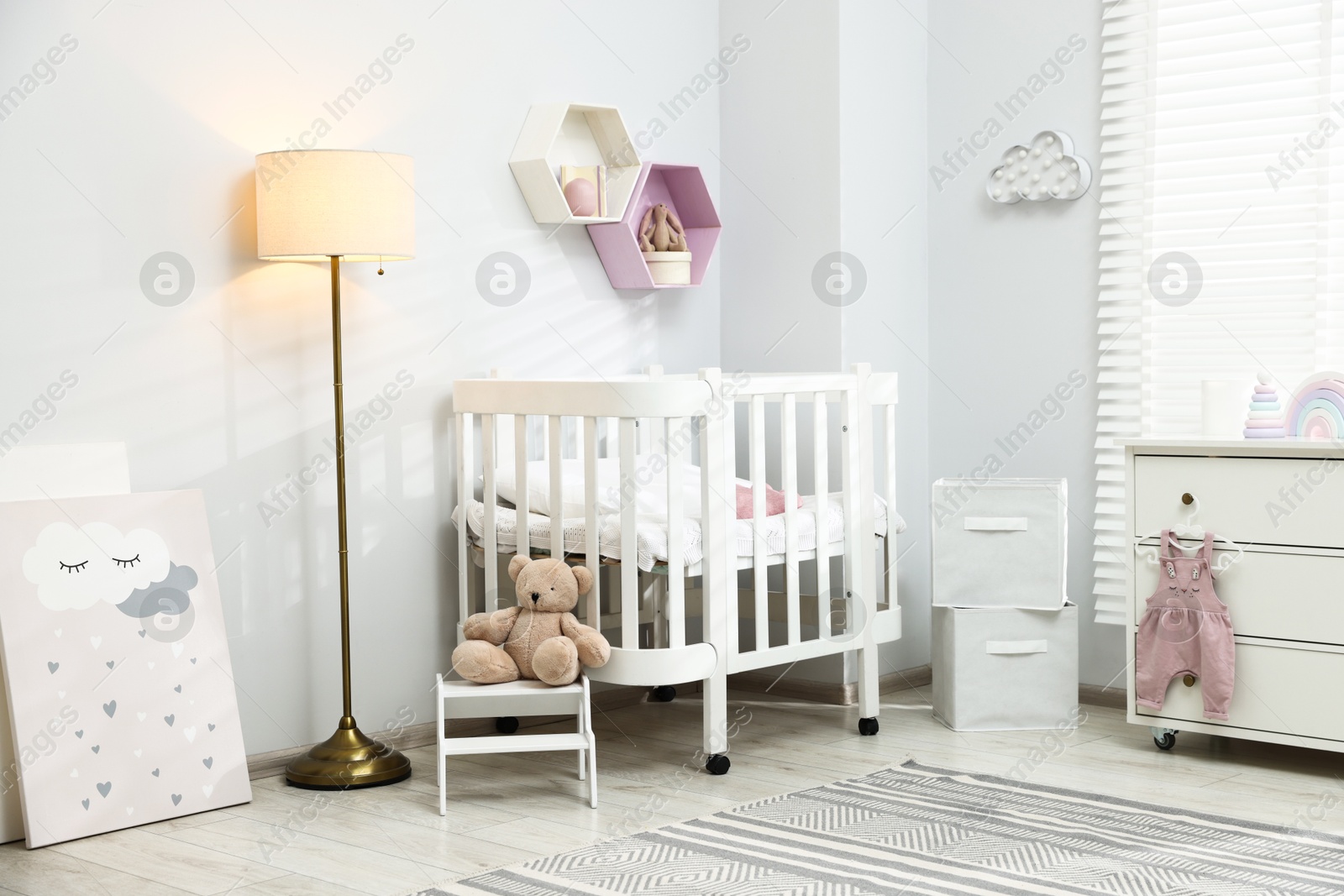 Stylish white crib, chest of drawers, lamp, toys and decor elements in baby's room. Interior design Photo of Stylish white crib, chest of drawers, lamp, toys and decor elements in baby's room. Interior design