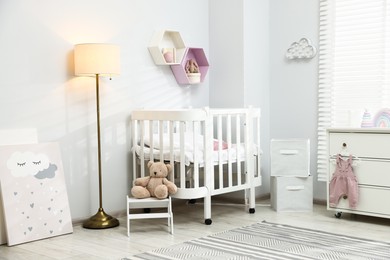 Photo of Stylish white crib, chest of drawers, lamp, toys and decor elements in baby's room. Interior design