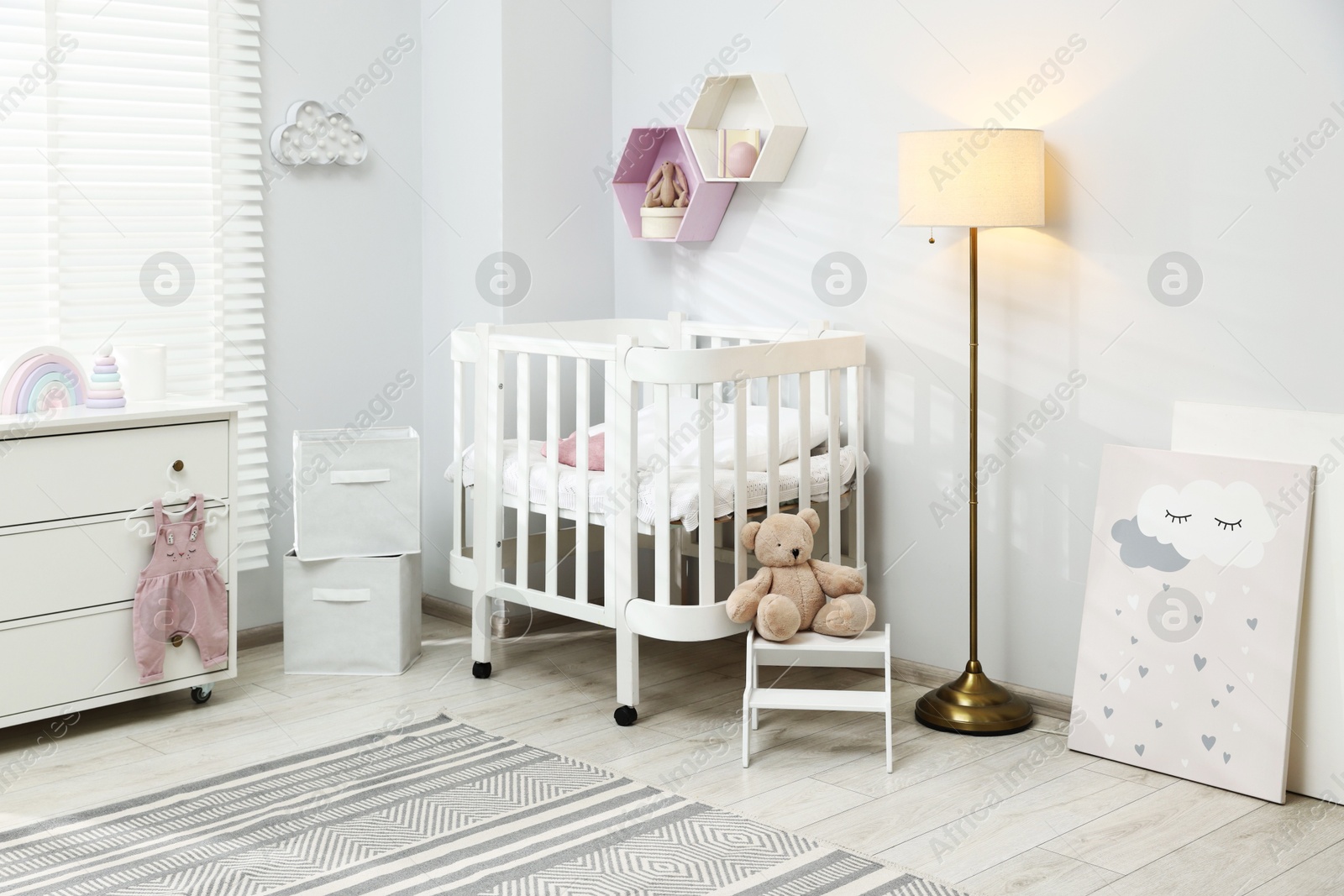 Stylish white crib, chest of drawers, lamp, toys and decor elements in baby's room. Interior design Photo of Stylish white crib, chest of drawers, lamp, toys and decor elements in baby's room. Interior design