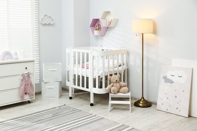 Photo of Stylish white crib, chest of drawers, lamp, toys and decor elements in baby's room. Interior design