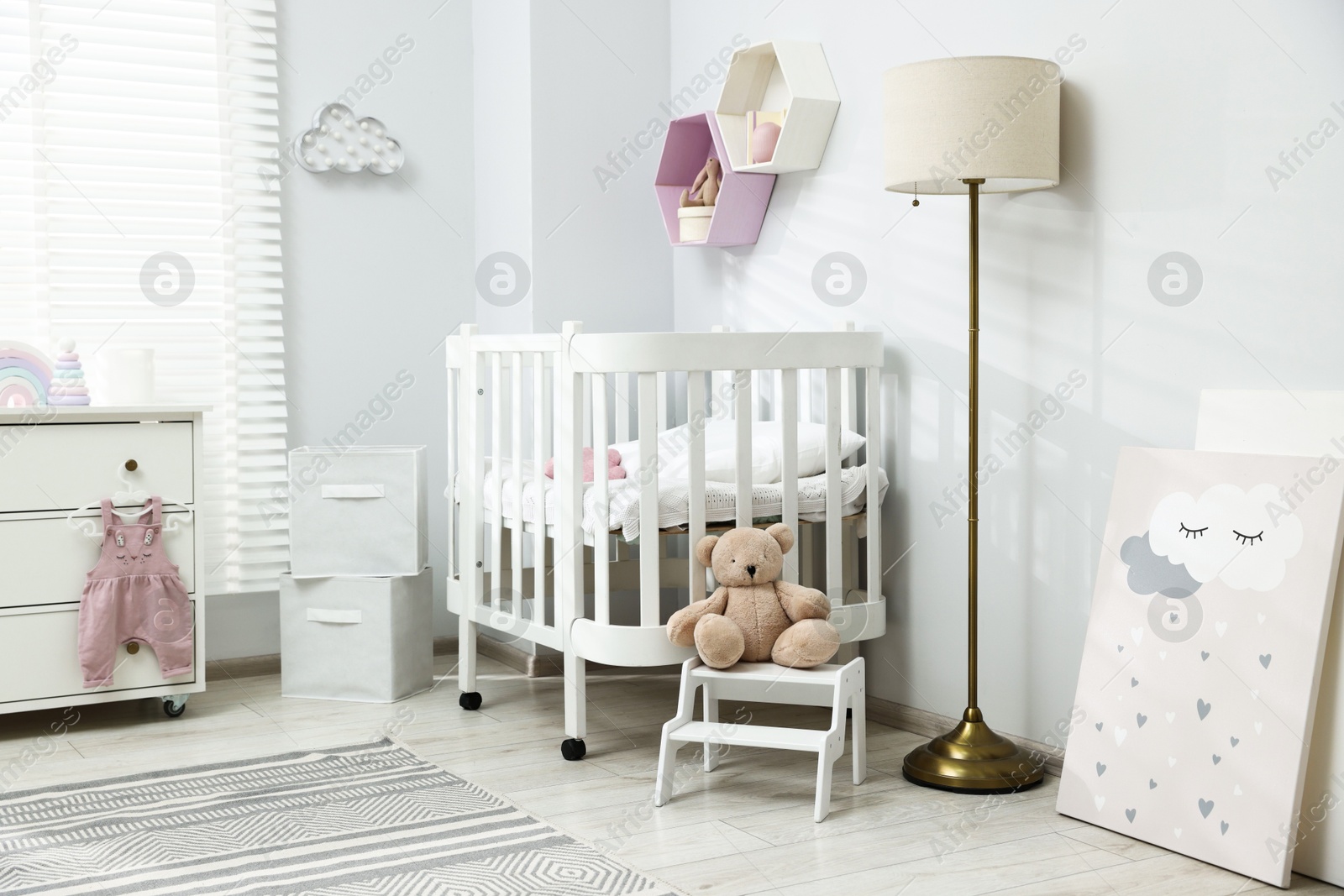 Stylish white crib, chest of drawers, lamp, toys and decor elements in baby's room. Interior design Photo of Stylish white crib, chest of drawers, lamp, toys and decor elements in baby's room. Interior design