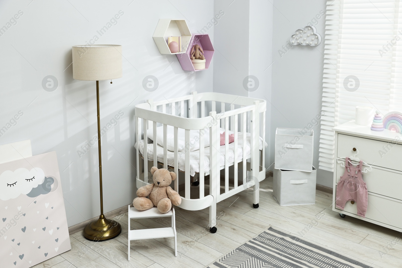 Stylish white crib, chest of drawers, lamp, toys and decor elements in baby's room. Interior design Photo of Stylish white crib, chest of drawers, lamp, toys and decor elements in baby's room. Interior design