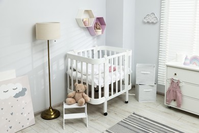 Photo of Stylish white crib, chest of drawers, lamp, toys and decor elements in baby's room. Interior design