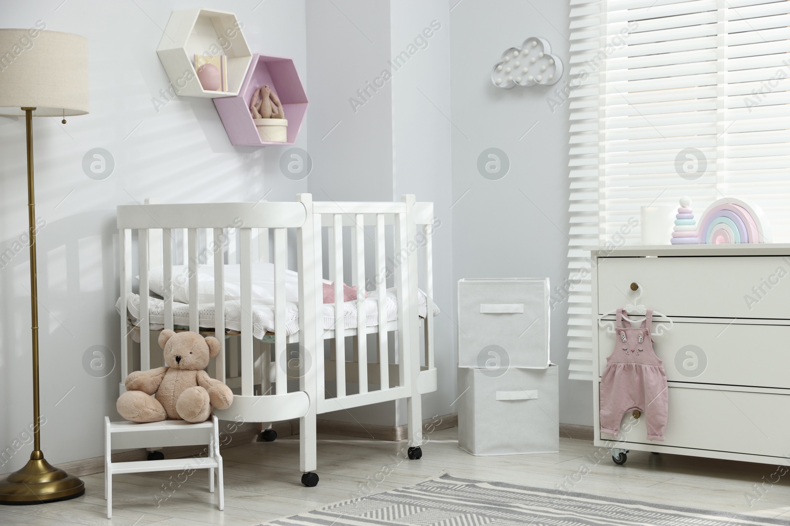 Stylish white crib, chest of drawers, lamp, toys and decor elements in baby's room. Interior design Photo of Stylish white crib, chest of drawers, lamp, toys and decor elements in baby's room. Interior design