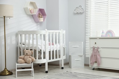 Photo of Stylish white crib, chest of drawers, lamp, toys and decor elements in baby's room. Interior design