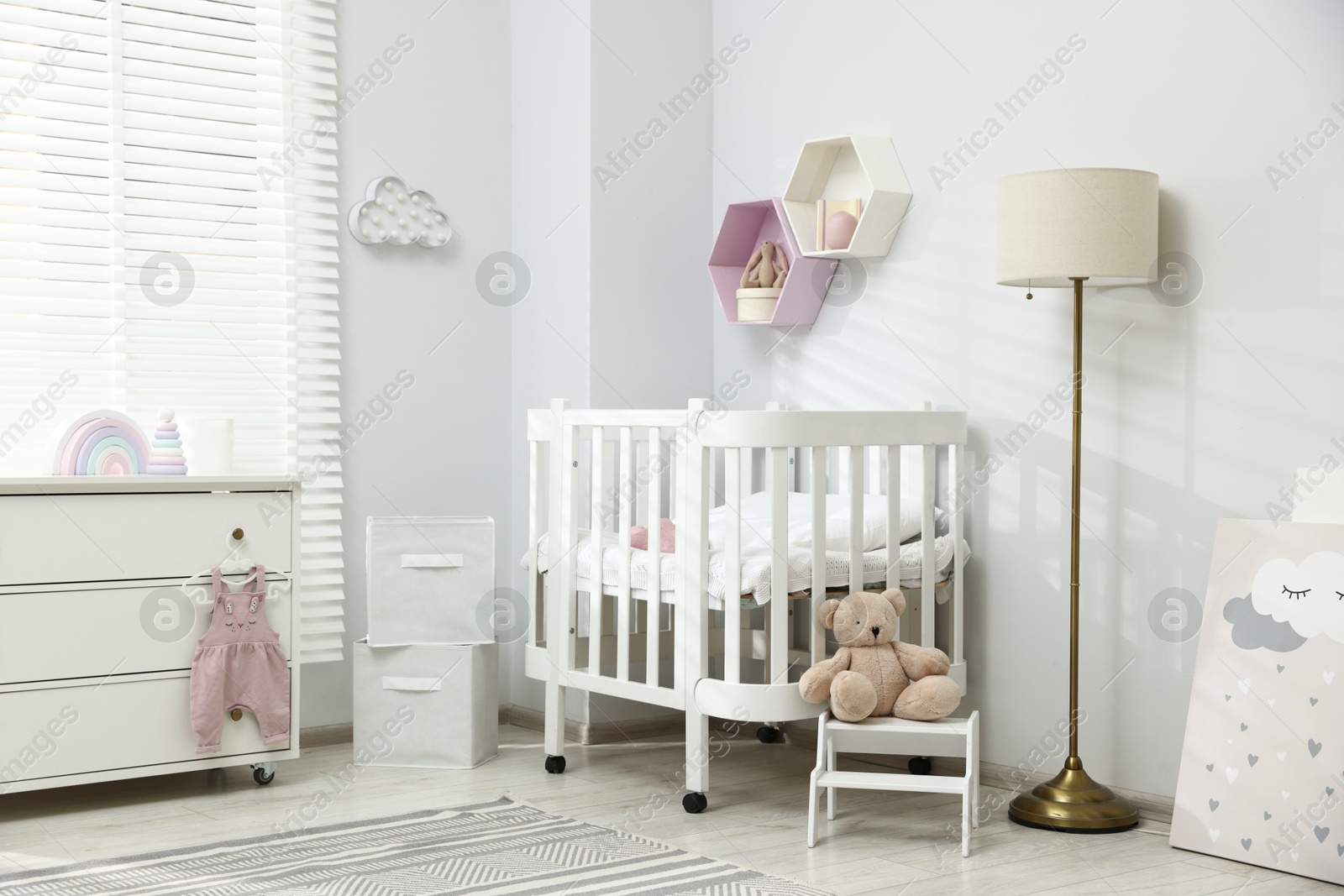 Stylish white crib, chest of drawers, lamp, toys and decor elements in baby's room. Interior design Photo of Stylish white crib, chest of drawers, lamp, toys and decor elements in baby's room. Interior design