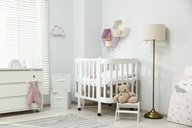 Photo of Stylish white crib, chest of drawers, lamp, toys and decor elements in baby's room. Interior design