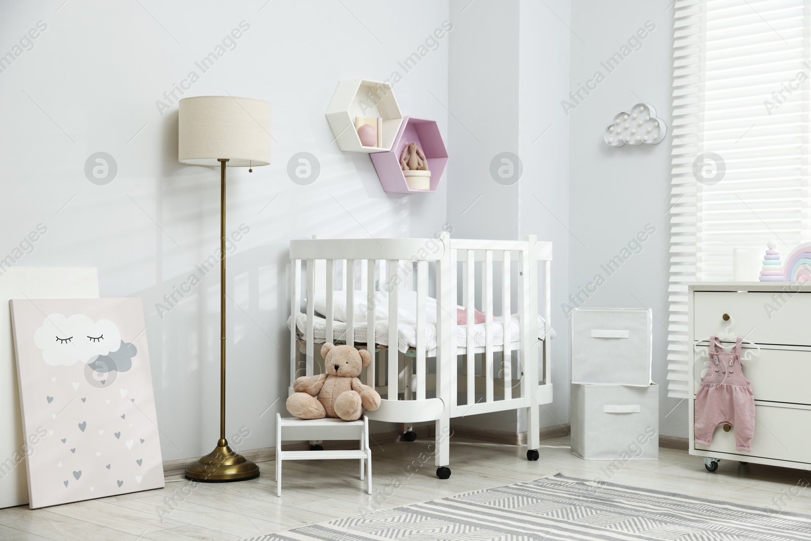 Stylish white crib, chest of drawers, lamp, toys and decor elements in baby's room. Interior design Photo of Stylish white crib, chest of drawers, lamp, toys and decor elements in baby's room. Interior design