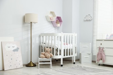 Photo of Stylish white crib, chest of drawers, lamp, toys and decor elements in baby's room. Interior design