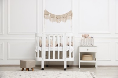 Photo of Comfortable crib, pouf, bedside table and toys near white wall in baby's room. Interior design