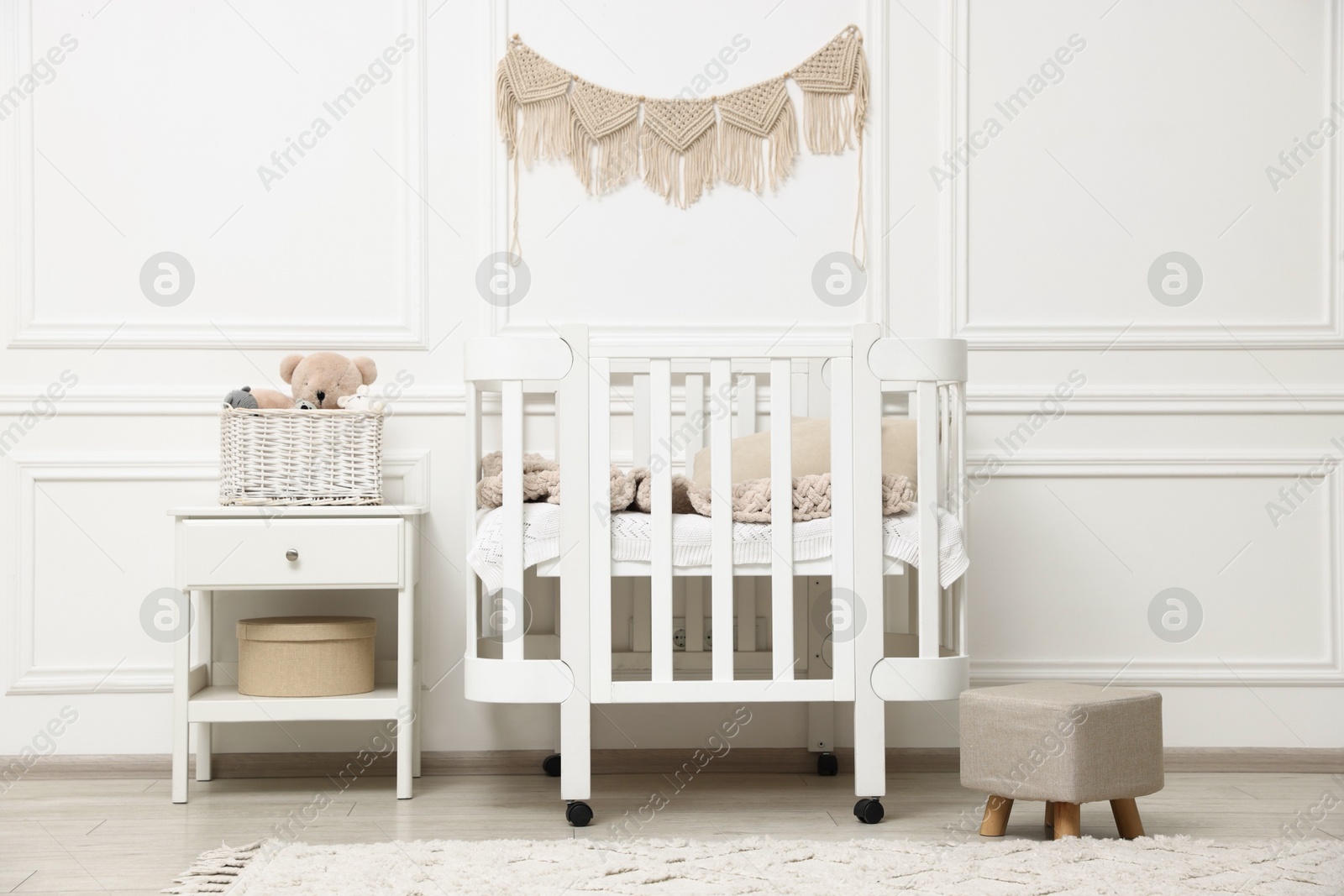 Comfortable crib, pouf, bedside table and toys near white wall in baby's room. Interior design Photo of Comfortable crib, pouf, bedside table and toys near white wall in baby's room. Interior design
