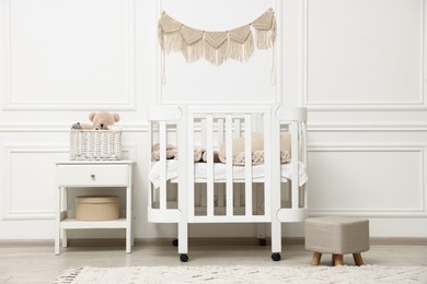 Photo of Comfortable crib, pouf, bedside table and toys near white wall in baby's room. Interior design