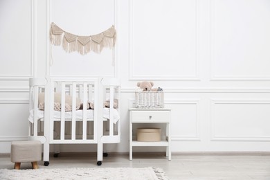 Photo of Comfortable crib, pouf, bedside table and toys near white wall in baby's room. Interior design