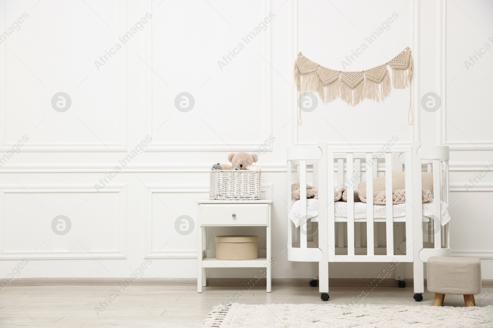 Comfortable crib, pouf, bedside table and toys near white wall in baby's room. Interior design Photo of Comfortable crib, pouf, bedside table and toys near white wall in baby's room. Interior design