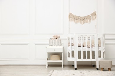 Photo of Comfortable crib, pouf, bedside table and toys near white wall in baby's room. Interior design