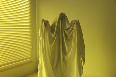 Photo of Creepy ghost. Man covered with white sheet indoors in yellow light