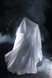 Photo of Creepy ghost. Man covered with white sheet in smoke on black background