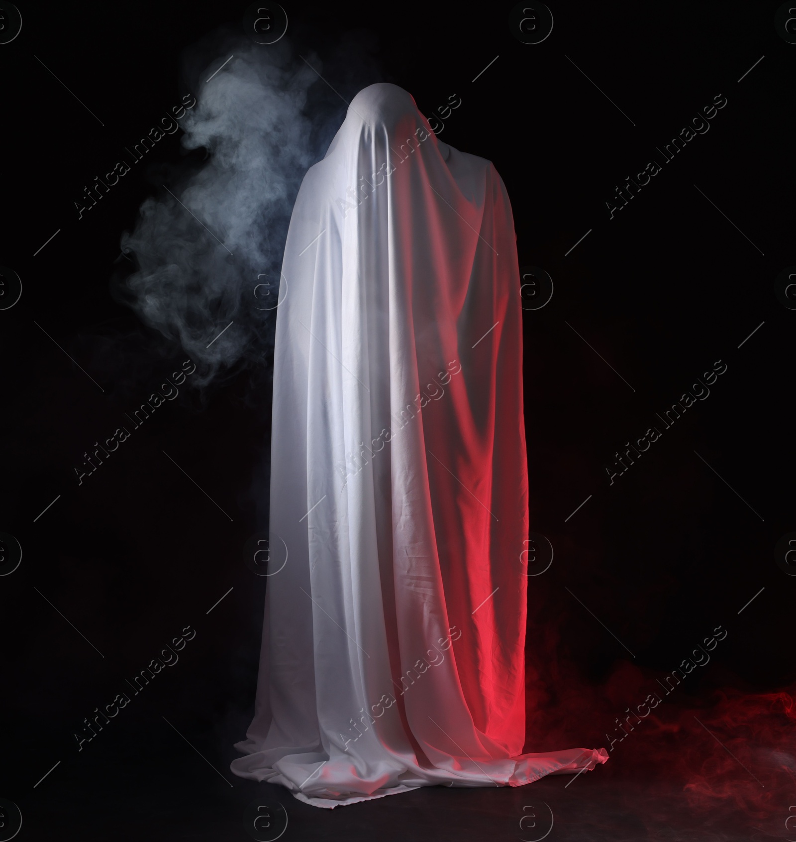Photo of Creepy ghost. Man covered with white sheet in smoke on black background