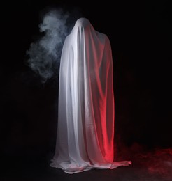 Photo of Creepy ghost. Man covered with white sheet in smoke on black background