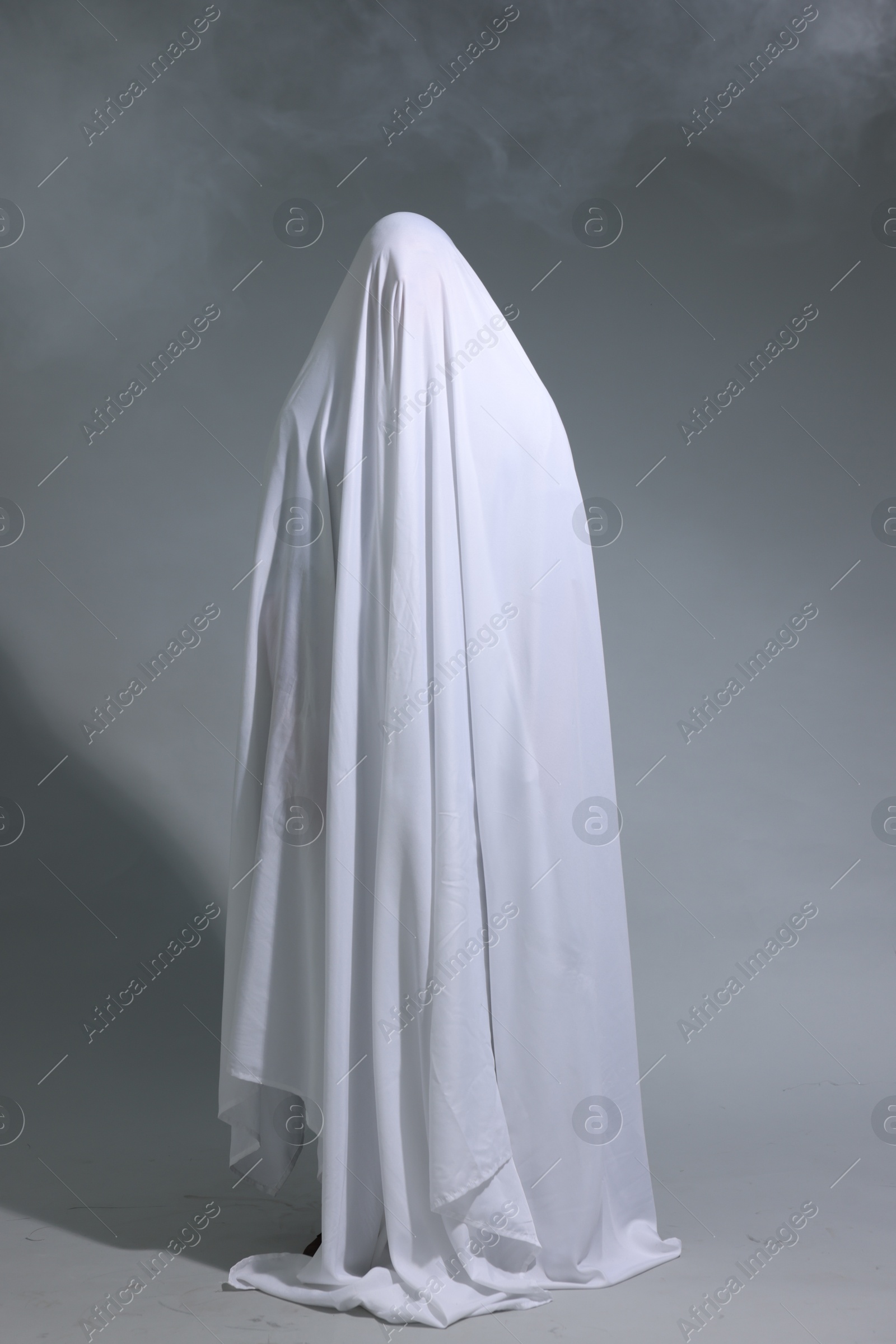 Creepy ghost. Man covered with white sheet in smoke on light grey background Photo of Creepy ghost. Man covered with white sheet in smoke on light grey background