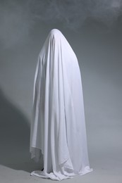 Photo of Creepy ghost. Man covered with white sheet in smoke on light grey background