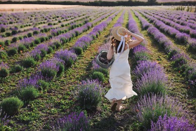 Photo of Woman with straw hat and basket of beautiful flowers running through lavender field, back view