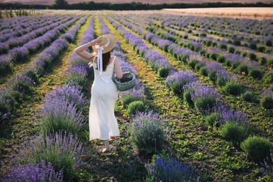 Photo of Woman with straw hat and basket of beautiful flowers running through lavender field, back view
