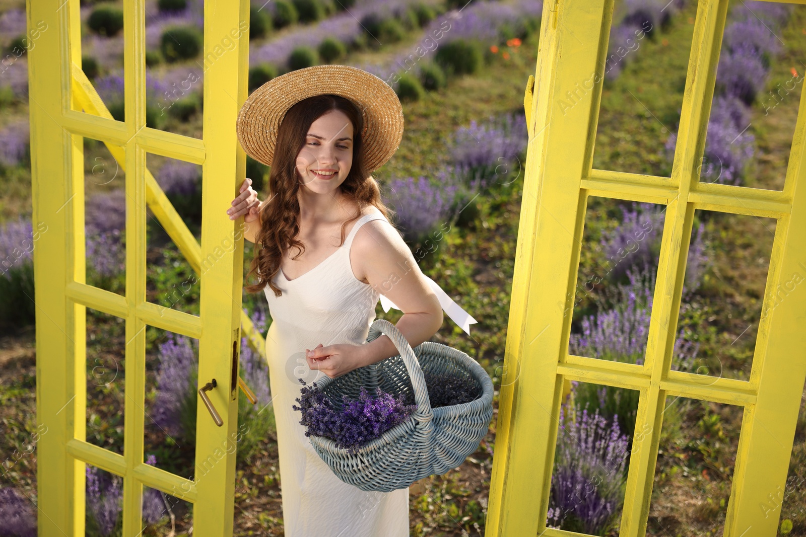Beautiful young woman with wicker basket of lavender flowers near yellow decorative door frame in field Photo of Beautiful young woman with wicker basket of lavender flowers near yellow decorative door frame in field