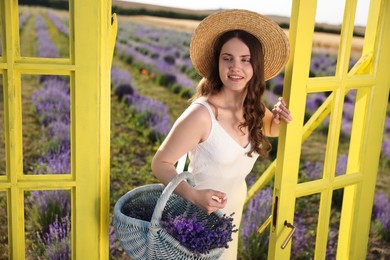 Photo of Beautiful young woman with wicker basket of lavender flowers near yellow decorative door frame in field