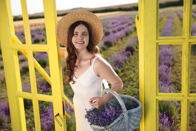 Photo of Beautiful young woman with wicker basket of lavender flowers near yellow decorative door frame in field