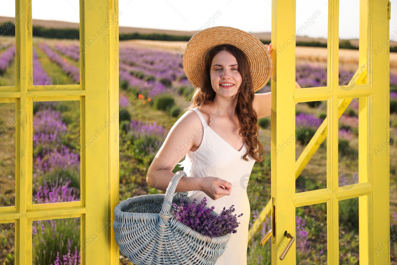 Beautiful young woman with wicker basket of lavender flowers near yellow decorative door frame in field Photo of Beautiful young woman with wicker basket of lavender flowers near yellow decorative door frame in field