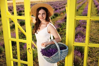 Photo of Beautiful young woman with wicker basket of lavender flowers near yellow decorative door frame in field