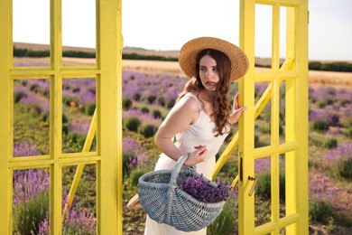 Photo of Beautiful young woman with wicker basket of lavender flowers near yellow decorative door frame in field