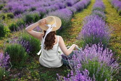 Photo of Woman with wicker basket of beautiful lavender flowers sitting in field, back view