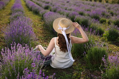Photo of Woman with wicker basket of beautiful lavender flowers sitting in field, back view
