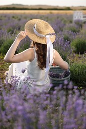 Photo of Woman with wicker basket of beautiful lavender flowers sitting in field, back view