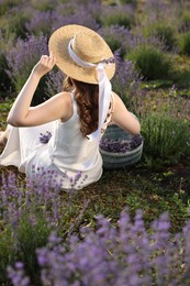 Photo of Woman with wicker basket of beautiful lavender flowers sitting in field, back view