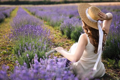 Photo of Woman with wicker basket of beautiful lavender flowers sitting in field, back view