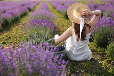 Photo of Woman with wicker basket of beautiful lavender flowers sitting in field, back view