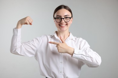 Photo of Strength. Happy businesswoman in glasses pointing at her biceps on grey background